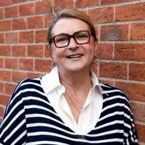 Rapid Transformational Therapist, Samantha Baker, stood against a brick wall wearing glasses, a white shirt, and a striped jumper - she's looking directly at the camera.