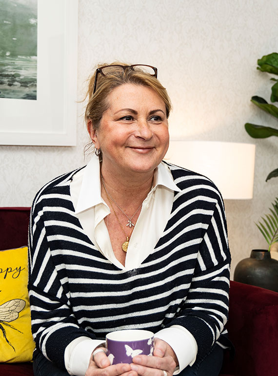 Samantha Baker - Rapid Transformational Therapist, Nottingham. Samantha sits on a deep red sofa with a cup of coffee. She has hear hair tied back, glasses on her head, and is wearing a white shirt with a striped jumper over.